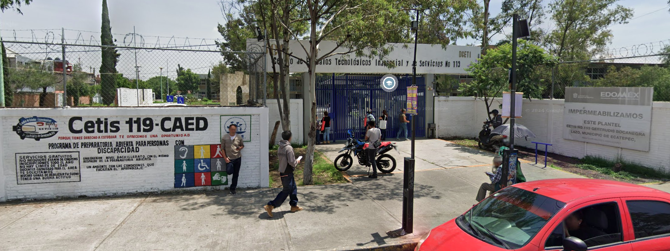 Fotografía en la que se muestra la entrada del plantel, conformada por una reja de color azul. Arriba de la reja hay un muro blanco en el que está escrito el nombre del plantel. Del lado izquierdo de la reja hay un muro de color blanco que tiene escritos los datos del plantel.